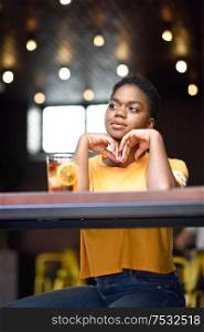 Young thoughtful black woman with very short hair taking a glass of cold tea in an urban cafe.. Young black woman with very short hair taking a glass of cold tea.