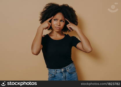 Young thoughtful mixed race woman with curly hair holds fingers on temples and trying to concentrate, demonstrating thinking process while posing isolated over beige wall. Concentration concept. Young thoughtful mixed race woman holds fingers on temples and trying to concentrate