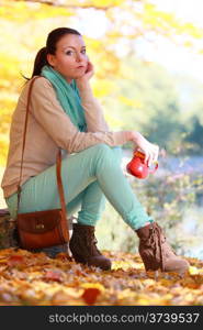 Young thoughtful pensive woman in the autumn park relaxing enjoying hot drink coffee or tea, holding red mug with warm beverage. Yellow leaves background