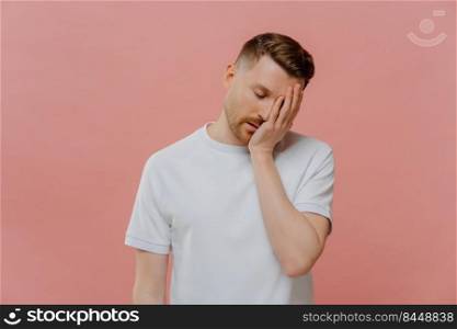 Young tired bearded man in white tshirt with closed eyes feeling sleepy, doing facepalm gesture while posing on pastel rose background, putting hand on his face and showing disappointment. Young exhausted caucasian man posing against pink background