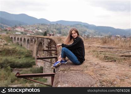 Young tourist girl sitting on viaduct with old railway tracks. Old railway viaduct in the mountain resort village of Vorokhta. Ukraine, Carpathians. Young tourist girl sitting on viaduct with old railway tracks. Old railway viaduct in the mountain resort village of Vorokhta. Ukraine, Carpathians.