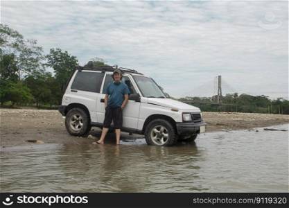 Young tourist man next to his all-terrain vehicle on the edge of a river in the Amazon jungle during a cloudy day. Young tourist man next to his all-terrain vehicle on the edge of a river in the Amazon jungle