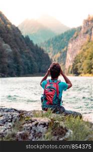 Young tourist with backpack looks through a binoculars on mountains peaks, stands on a rock over a river. Boy spends a vacation in mountains, wandering with backpack, he is wearing sports summer clothes