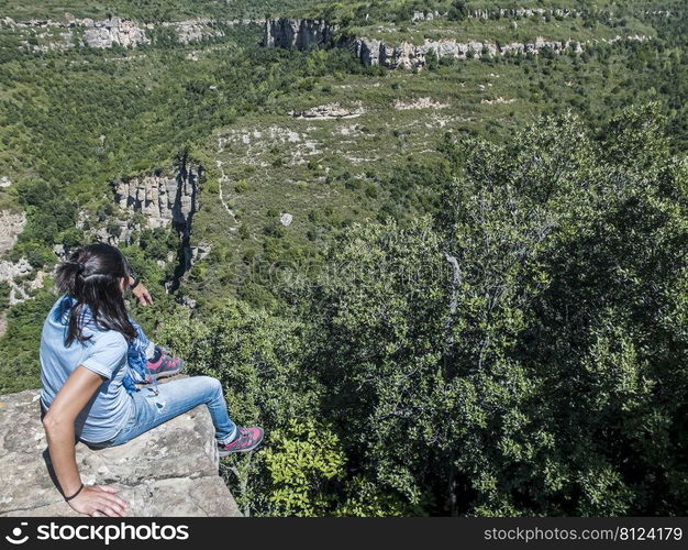 Young traveling woman sitting on the top of the mountain cliff with relaxing mood and watching beautiful view of woods and blue sky and clouds on vacation.