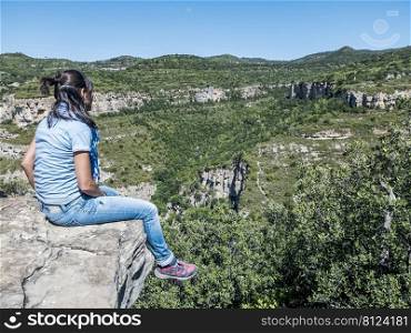 Young traveling woman sitting on the top of the mountain cliff with relaxing mood and watching beautiful view of woods and blue sky and clouds on vacation.