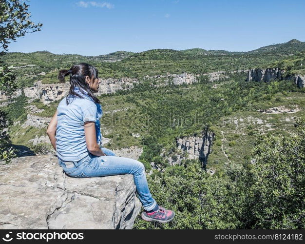 Young traveling woman sitting on the top of the mountain cliff with relaxing mood and watching beautiful view of woods and blue sky and clouds on vacation.