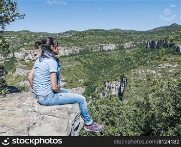 Young traveling woman sitting on the top of the mountain cliff with relaxing mood and watching beautiful view of woods and blue sky and clouds on vacation.