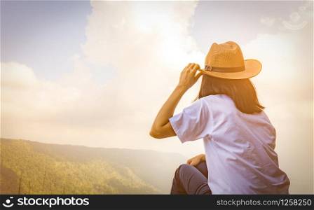 Young traveling woman wearing hat and sitting on the top of the mountain cliff with relaxing mood and watching beautiful view of woods and blue sky and clouds on vacation. Asian woman travel alone.