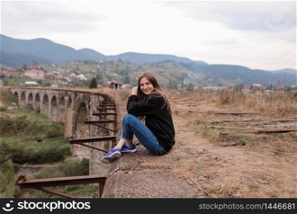 Young traveling woman with backpack sitting high on the top of the mountain with waving hair, evening with sunset. Young tourist girl sitting on viaduct with old railway tracks.. Young traveling woman with backpack sitting high on the top of the mountain with waving hair, evening with sunset.