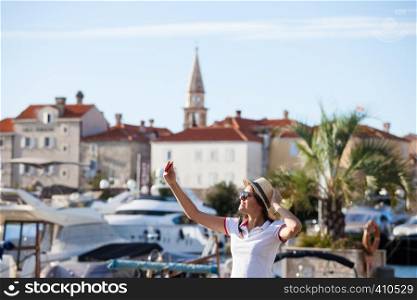 Young travelling woman in hat taking selfie photos over summer sunny mediterranean city with smartphone camera. Brunette girl making photography on summer vacation