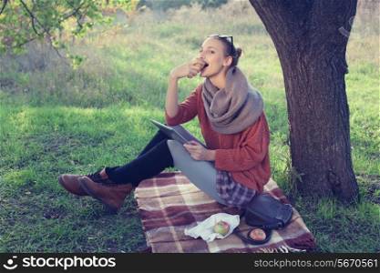 Young trendy woman eating apple and using tablet computer during picnic