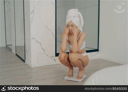 Young unhappy slender caucasian girl squatting barefoot on electronic scales with sad face on hands in stylish bedroom interior, female could not lose weight during dieting. Young unhappy caucasian girl sitting barefoot on electronic scales
