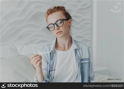 Young unhealthy red-haired lady holding thermometer while sitting on bed at home, feeling sick, suffering from flu influenza, ill woman measuring temperature in morning, female having covid19 symptoms. Young unhealthy red-haired lady holding thermometer while sitting on bed at home