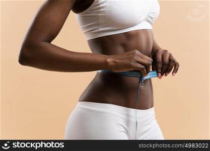 Young unrecognizable african woman measuring her waist in centimeters on beige background