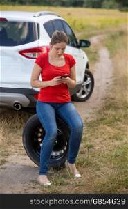 Young upset woman siting on spare tire next to broken car at field and searching for service phone number
