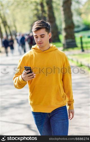 Young urban man using smartphone walking in street in an urban park in London, UK.. Young urban man using smartphone walking in street in an urban park in London.