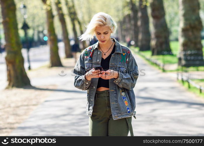 Young urban woman with modern hairstyle using smartphone walking in street in an urban park in London, UK.. Young urban woman with modern hairstyle using smartphone walking in street in an urban park.