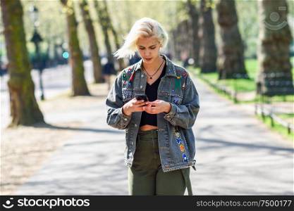 Young urban woman with modern hairstyle using smartphone walking in street in an urban park in London, UK.. Young urban woman with modern hairstyle using smartphone walking in street in an urban park.