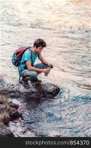 Young wanderer takes pure water from a river into mug. He is sitting on a rock over the river, rests during a hike, spends a vacation on wandering with backpack, he is wearing sport summer clothes