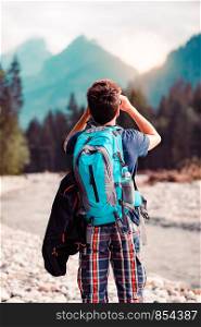 Young wanderer with backpack looks through a binoculars on mountains peaks, stands over a river. Boy spends a vacation in mountains, wandering with backpack, he is wearing sports summer clothes