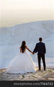 Young wedding couple stands over a cliff holding hands and looks into the distance. Man and woman in wedding attire. Lovers stand on the background of white Pamukkale travertines. The view from the back. Concept of love and relationship. Young wedding couple stands over cliff holding hands and looks into the distance