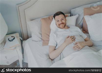 Young well rested man in white tee shirt waking up in morning with smile, happy after sleeping enough, lying in comfortable bed while covered with blankets, lots of different pillows. Young well rested man waking up in morning at home