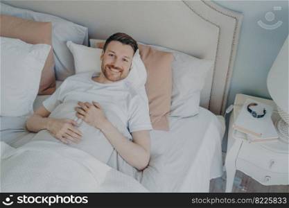 Young well rested man in white tee shirt waking up in morning with smile, happy after sleeping enough, lying  in comfortable bed while covered with blankets, lots of different pillows. Young well rested man waking up in morning at home