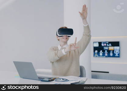 Young white woman in VR goggles clicks virtual buttons. Girl is sitting at the desk in front of laptop in office. Happy businesswoman working on project in cyberspace. Concept of innovation.. Young white woman in VR goggles working on project in cyberspace in front of laptop in office.