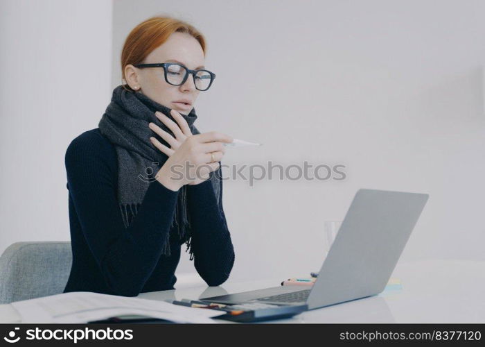 Young white woman is sick. Unwell lady girl is working in office in scarf and looking at thermometer. Business employee feels ill and has fever and temperature sitting at the desk.. Young white woman is sick while working in office in scarf and looking at thermometer.