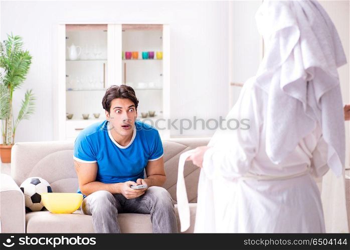 Young wife is trying to seduce husband from watching football an. Young wife is trying to seduce husband from watching football and tv. Young wife is trying to seduce husband from watching football an