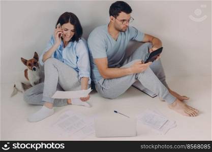Young woman and man sit back to each other, do accounts together, make necessary calculations, surrounded with paper documents, sit on floor in empty room with dog. People and finances concept