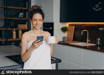 Young woman applies anti wrinkle eye patches and relaxing at home texting on phone. Happy hispanic girl wrapped in towel after spa procedures. Body care, beauty and wellness. Modern interior.. Young woman applies anti wrinkle eye patches and relaxing at home texting on phone.