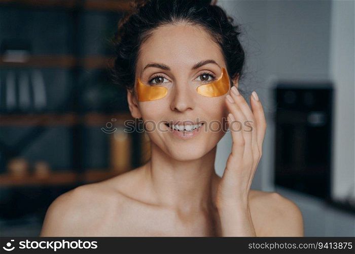 Young woman applies collagen eye patches, happy European girl in spa mode. Golden anti-wrinkle patches enhance skincare routine.