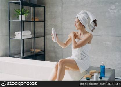 Young woman applies cream to her shoulder. Body lotion applying. Attractive caucasian girl wrapped in towel after bathing. Cosmetic products for smooth silky skin. Spa procedure at home.. Young woman applies cream to her shoulder. Cosmetic products and spa procedure for silky skin.