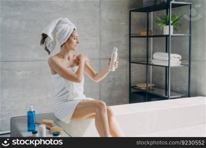Young woman applies cream to her shoulder. Body lotion applying. Attractive caucasian girl wrapped in towel after bathing. Cosmetic products for smooth silky skin. Spa procedure at home.. Young woman applies cream to her shoulder. Cosmetic products and spa procedure for silky skin.
