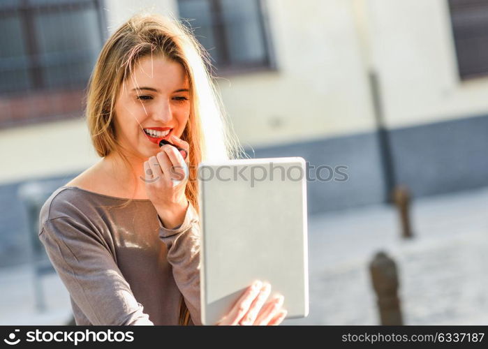 Young woman applying lipstick looking at tablet in urban background