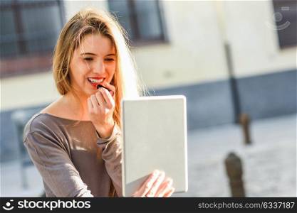 Young woman applying lipstick looking at tablet in urban background