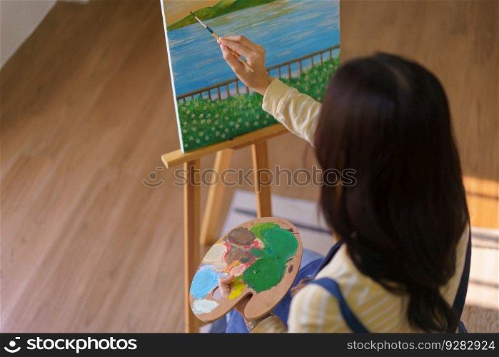 Young woman artist holding color palette and painting picture in canvas on easel at art studio.