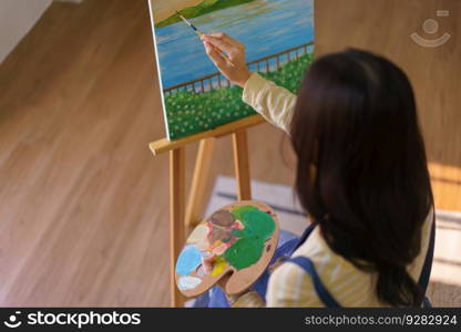 Young woman artist holding color palette and painting picture in canvas on easel at art studio.