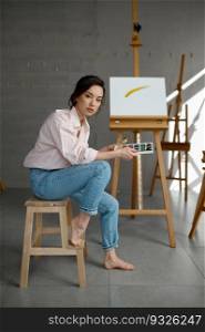 Young woman artist with creative accessories sitting on chair at artwork workplace with wooden easel on background. Young woman artist sitting on chair at home artwork workplace