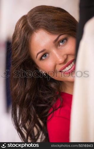 Young woman at clothes shop peeping through clothes smiling happy and excited at camera.