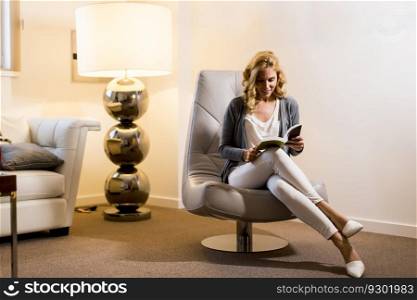 Young woman at home sitting on modern chair in front of modern l& relaxing in her living room reading book