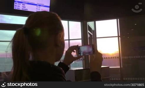 Young woman at the airport using smart phone to take pictures of sunset through the window