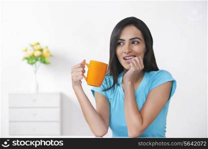 Young woman biting nail while holding coffee cup at home