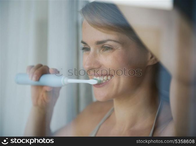 Young woman brushing teeth with electric toothbrush and looking in window