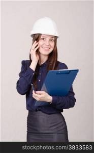 Young woman builder in helmet standing with a folder in hand and talking on phone