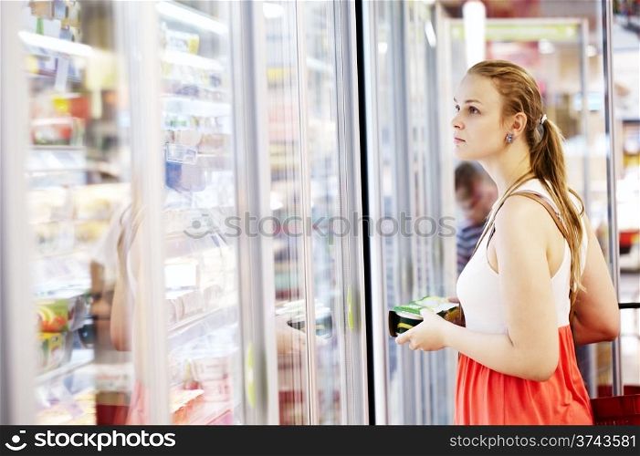 Young woman buying dairy or refrigerated groceries at the supermarket in the refrigerated section opening glass door of the fridge
