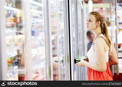 Young woman buying dairy or refrigerated groceries at the supermarket in the refrigerated section opening glass door of the fridge