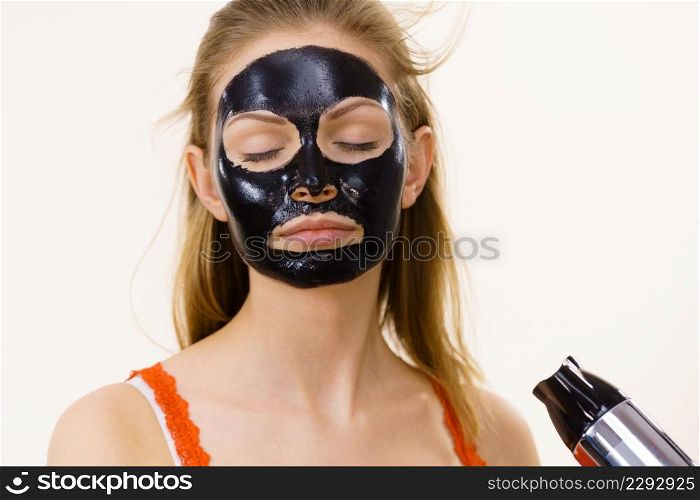 Young woman carbo black peel-off mask on her face, girl drying cosmetic to removing it, using hair dryer. Beauty treatment. Skin care.. Girl drying peel-off black mask on face