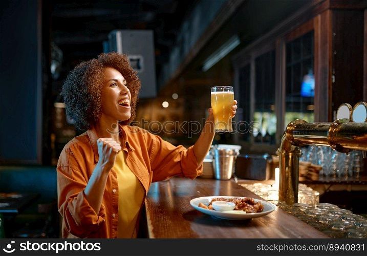 Young woman cheering for favorite team watching match in sports bar. Cheerful excited millennial girl sport fan drinking beer while sitting at counter desk. Young woman cheering for favorite team watching match in sports bar